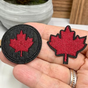 Peut inclure: Deux écussons brodés représentant une feuille d'érable rouge. L'un est rond avec un fond noir, l'autre a la forme d'une feuille d'érable avec un bord noir. Les écussons sont tenus dans une main.