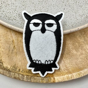 May include: Black and white embroidered owl patch with a white fluffy body and black eyes. The owl is sitting on a wooden surface.