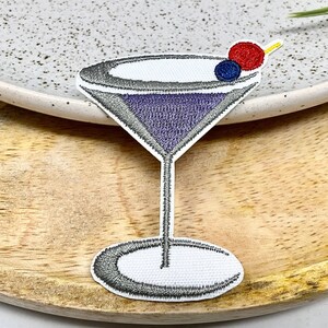 Peut inclure: Un patch brodé thermocollant représentant un verre à martini violet avec une cerise rouge et bleue sur le dessus. Le patch est sur une surface en bois.