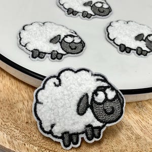 May include: Four white sheep patches with black outlines and details. The sheep have fluffy white bodies, black faces, and black legs. One patch is on a wooden surface, and the other three are on a white plate.