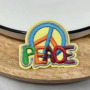 May include: A colourful embroidered patch featuring a peace sign and the word "peace" in rainbow colours. The patch is on a light brown wooden surface.