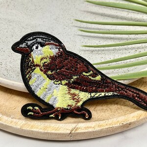 Peut inclure: Un patch brodé représentant un oiseau brun et jaune avec des points de couture noirs. L'oiseau est perché sur une surface en bois.