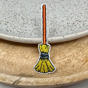May include: Embroidered patch of a yellow and black broom with an orange handle. The patch is on a white background and is placed on a wooden tray with a white speckled plate.