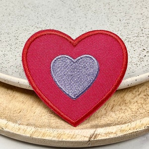 Peut inclure: Un patch en forme de cœur rouge avec un plus petit cœur violet à l'intérieur. Le patch est sur une surface en bois.