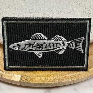 May include: A black and white embroidered patch of a fish with a grey border. The fish is white with black details and is on a black background.