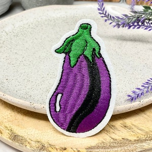 May include: A purple and green embroidered eggplant patch. The eggplant is outlined in black thread and has a green stem.