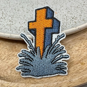 Op de afbeelding: Een geborduurde strijk-patch met een blauw kruis met een oranje centrum, omringd door gestileerd blauw en wit gras.