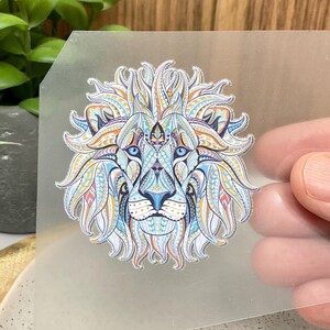 Op de afbeelding: Een transparant vel met een kleurrijk, gepatroneerd leeuwenontwerp. De leeuwenmanen is blauw, oranje en wit. Het gezicht van de leeuw is wit met blauwe ogen en een gele neus.