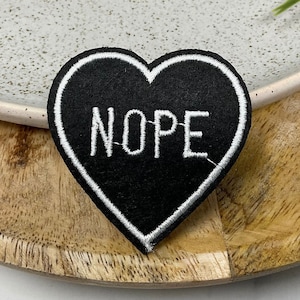 Puede incluir: Parche negro en forma de corazón con contorno blanco y la palabra "NOPE" bordada en blanco.