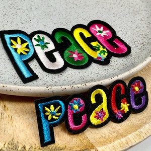 May include: Two embroidered patches spelling out "Peace" in colourful letters with flower designs. The patches are on a wooden surface.
