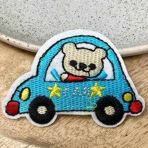 Peut inclure: Patch thermocollant brodé représentant un ours en dessin animé conduisant une voiture bleue avec des étoiles jaunes et le mot "CAR" sur le côté.
