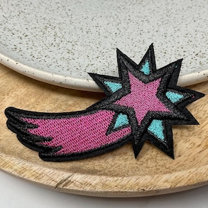 May include: A pink and blue shooting star with a black outline. The star is a five-pointed star with a black outline and a pink centre. The shooting star is on a wooden surface.