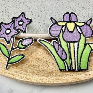 Peut inclure: Deux patchs brodés thermocollants représentant des fleurs violettes. Un patch est une seule fleur avec des feuilles vertes et un centre jaune. L'autre patch est un groupe de trois fleurs avec des feuilles vertes.