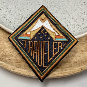 May include: A diamond-shaped embroidered patch with a dark blue background, gold and orange accents, and the word "TRAVELER" in gold lettering. The patch features a stylised image of a tent with a crescent moon and stars.