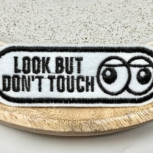 Peut inclure: Un écusson en feutre blanc avec du texte brodé en noir et un dessin d'yeux de dessin animé. Le texte dit "LOOK BUT DON'T TOUCH". L'écusson est de forme ovale avec une bordure noire.