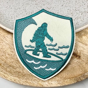 May include: A teal blue embroidered patch in the shape of a shield with a white background. The patch features a teal blue silhouette of a Bigfoot surfing on a wave.
