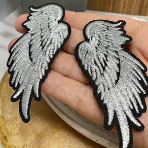 May include: Two silver angel wings with black outlines. The wings are embroidered and have a detailed design.