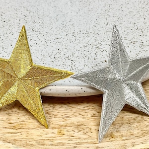 Può includere: Due stelle ricamate, una dorata e una argentata, su una superficie di legno.