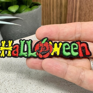 May include: An embroidered iron-on patch with the word "Halloween" in green, yellow, and red letters. A small red pumpkin with a jack-o-lantern face is in the centre of the word.