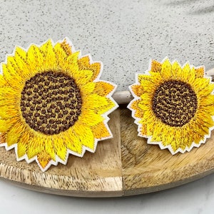 Peut inclure: Deux patchs brodés en forme de tournesol, l'un plus grand que l'autre, avec des pétales jaunes et des centres marrons. Les patchs sont sur une surface en bois.