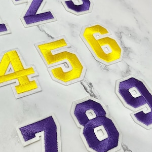 Peut inclure: Écussons de chiffres brodés en violet et or avec des bordures blanches. Les chiffres 1 à 9 sont visibles, avec un mélange de fil violet et or. Les écussons sont probablement destinés aux maillots de sport ou à d'autres vêtements.