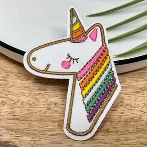 May include: Embroidered unicorn patch with a white body, a rainbow mane, and a gold-coloured outline. The unicorn has a pink ear and a pink dot for a cheek. The horn is striped with orange, yellow, and green.