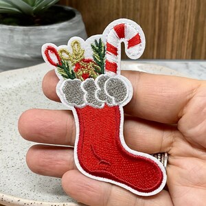 Peut inclure: Chaussette de Noël rouge et blanche avec une canne en sucre, des feuilles vertes et une boule dorée. La chaussette est brodée de bordures blanches et d'un revers blanc moelleux.