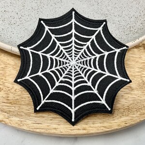 Pode incluir: Patch bordado de teia de aranha preto e branco. A teia é branca e o fundo é preto.