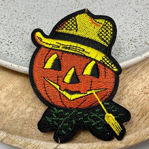 May include: A black and orange embroidered patch of a jack-o'-lantern wearing a yellow straw hat with a green stem and leaves.