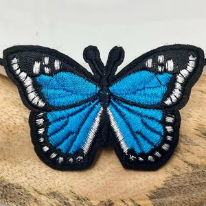 Peut inclure: Écusson papillon brodé avec une bordure noire et des ailes bleu vif. Le papillon a des détails blancs et est posé sur une surface en bois. Ce patch décoratif peut être repassé sur des vêtements ou des accessoires.