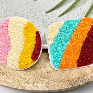 May include: Two colourful, round and square shaped embroidered patches with a striped design. The patches are made of a fluffy, textured fabric and feature a variety of colours, including red, orange, yellow, blue, and white.