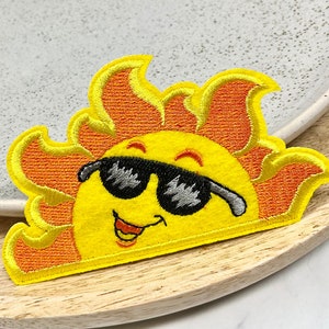 Peut inclure: Un soleil jaune avec un visage souriant portant des lunettes de soleil noires et des rayons orange. Le soleil est brodé sur un fond blanc.
