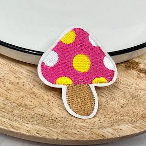 Pode incluir: Patch bordado de cogumelo rosa e branco com bolinhas amarelas. O cogumelo tem um caule castanho.