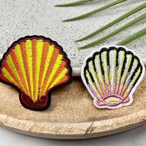 Peut inclure: Deux patchs en tissu brodés en forme de coquillages. L'un est jaune et orange avec un contour rouge. L'autre est blanc avec un centre rose et jaune et un contour noir.