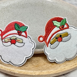 Peut inclure: Deux décorations de Noël en feutre brodées représentant le Père Noël avec une barbe blanche et un chapeau rouge. Chaque décoration est surmontée d'une branche de houx verte avec des baies rouges.