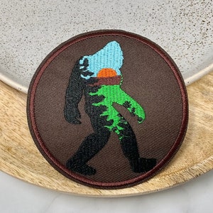 Op de afbeelding: Ronde geborduurde patch met een Bigfoot-ontwerp. De patch heeft een bruine achtergrond met een Bigfoot-silhouet gevuld met blauwe, oranje en groene kleuren. De patch is omrand met een donkerrode rand.