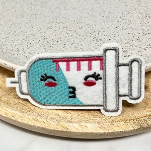 Peut inclure: Un patch brodé blanc et bleu représentant une seringue avec un visage de dessin animé mignon. La seringue a une aiguille rose et rouge et un œil qui cligne des deux côtés.