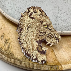 Peut inclure: Un patch brodé représentant une tête de lion en brun et beige avec un œil jaune. Le patch est sur une surface en bois.