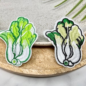 May include: Two embroidered iron-on patches of bok choy, one with green leaves and one with yellow leaves. Both patches have white stems and are on a wooden surface.