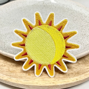 May include: A yellow embroidered sun patch with red rays and a white outline. The sun is partially eclipsed by a crescent moon.