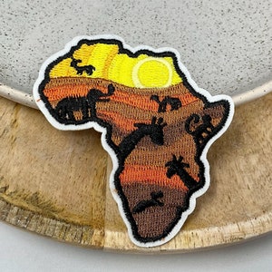 Könnte beinhalten: Ein gestickter Aufnäher in Form des afrikanischen Kontinents mit einer Sonnenuntergangsszene und verschiedenen Tieren. Der Aufnäher ist braun, orange und gelb.