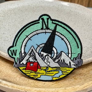 Peut inclure: Patch brodé avec un motif de boussole représentant une chaîne de montagnes, une tente et une aiguille de boussole pointant vers le nord. Le cadran de la boussole est bleu et blanc, avec un anneau extérieur vert. Les lettres "W" et "N" sont visibles sur la boussole.