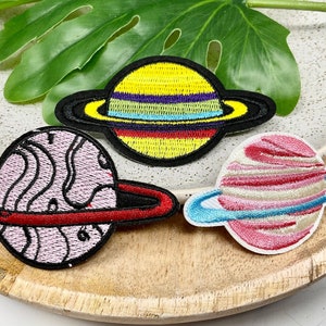 May include: Three embroidered planet patches. One planet is yellow with colorful stripes and a black ring. Another is pink with a red ring. The third is pink and white with a blue ring. All have black borders.