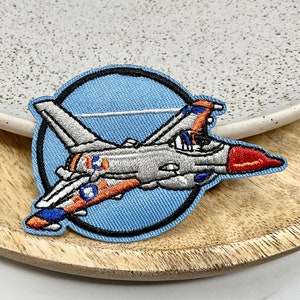 May include: Embroidered patch of a fighter jet in orange, blue and grey colours against a blue background. The patch is circular with a black border.