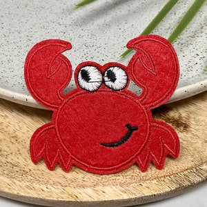 Op de afbeelding: Rode viltkrabben-applicatie met witte ogen en een zwarte glimlach. De krab zit op een houten oppervlak.