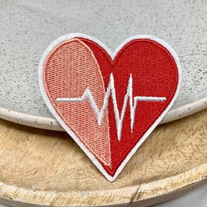 Peut inclure: Patch brodé en forme de cœur rouge et rose avec une ligne ECG blanche. Le patch est sur une surface en bois.