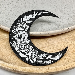 May include: Black embroidered patch of a crescent moon with white floral details. The moon is surrounded by white flowers and leaves.