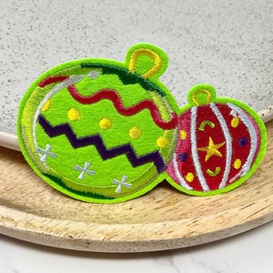 Peut inclure: Deux décorations de Noël brodées, l'une verte avec un motif en zigzag et des points jaunes, et l'autre rouge avec des rayures blanches et une étoile jaune.