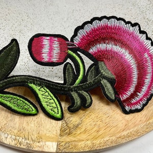 Peut inclure: Un patch brodé en forme de fleur rose et blanche avec des feuilles vertes et une tige. La fleur est un œillet stylisé avec un bord festonné.