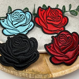 Peut inclure: Quatre écussons brodés en forme de rose, de couleurs bleu clair, rouge, noir et rouge foncé. Chaque écusson présente un motif de rose détaillé avec une bordure noire. Les écussons sont présentés sur une assiette mouchetée avec de la verdure.